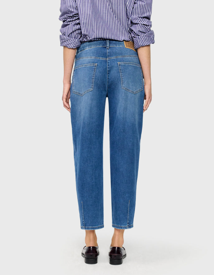 A model wearing a pair of medium-wash blue barrel jeans with a subtle vertical fade, paired with a purple and white striped shirt and dark brown loafers, against a light grey background.