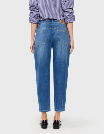 A model wearing a pair of medium-wash blue barrel jeans with a subtle vertical fade, paired with a purple and white striped shirt and dark brown loafers, against a light grey background.