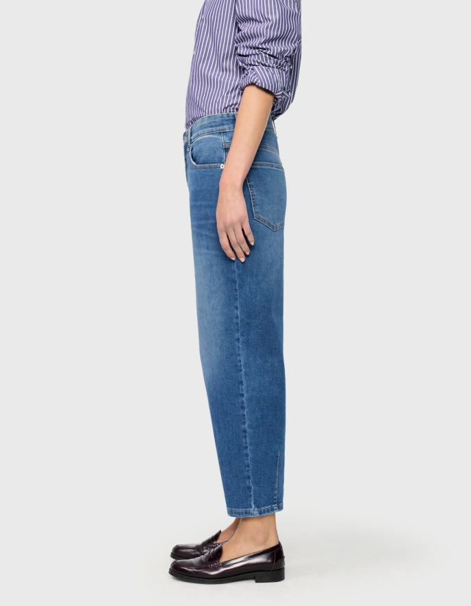 A model wearing a pair of medium-wash blue barrel jeans with a subtle vertical fade, paired with a purple and white striped shirt and dark brown loafers, against a light grey background.
