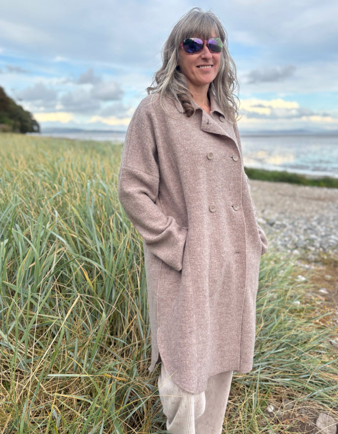 Woman wearing a beige boiled wool coat and sunglasses standing on a grassy beach.