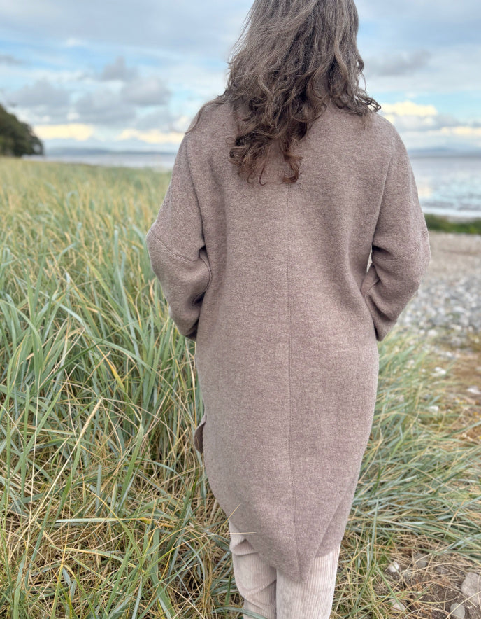 Woman wearing a beige boiled wool coat and sunglasses standing on a grassy beach.
