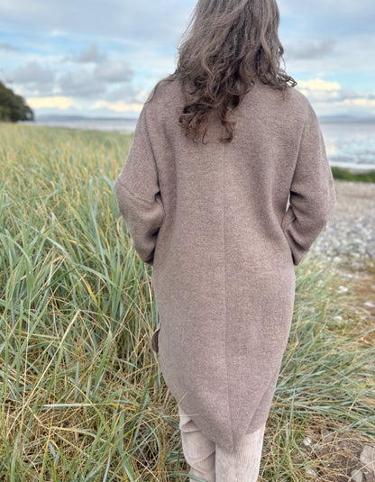 Woman wearing a beige boiled wool coat and sunglasses standing on a grassy beach.