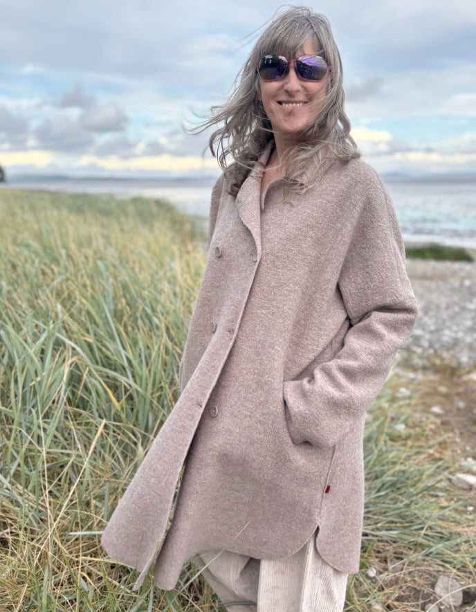 Woman wearing a beige boiled wool coat and sunglasses standing on a grassy beach.