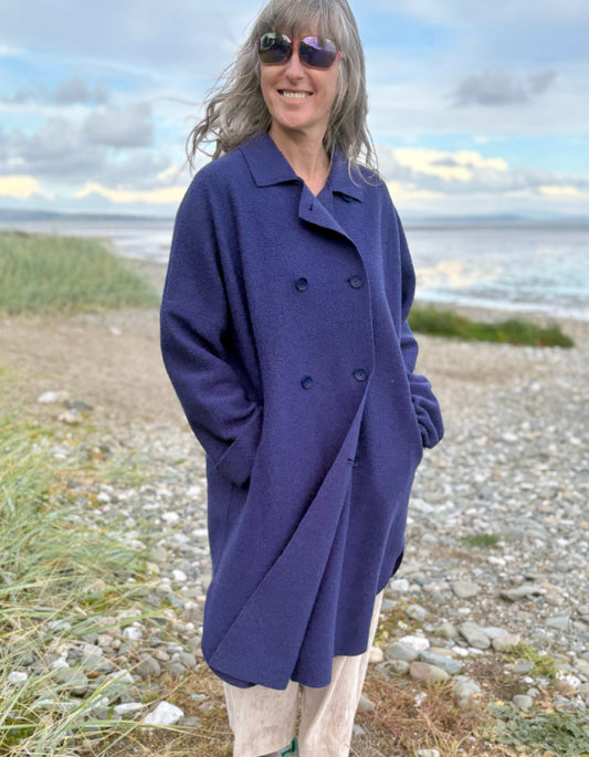 Person wearing a purple blue boiled wool coat standing on a beach with ocean and sky in the background