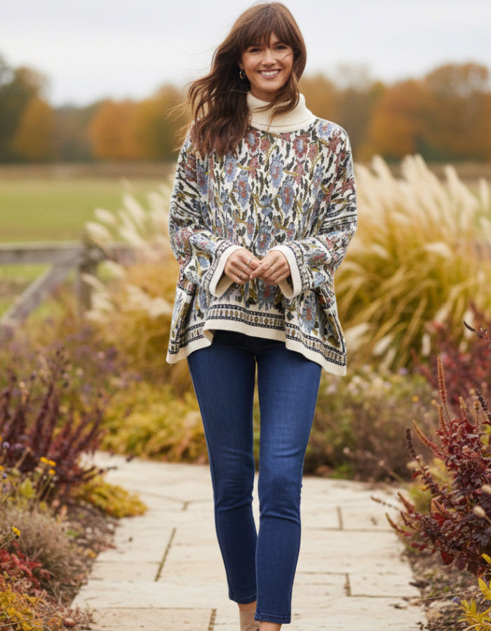 A woman stands on a stone path in a garden wearing a cream turtleneck sweater with a multicolor floral and geometric pattern, paired with dark blue skinny jeans.