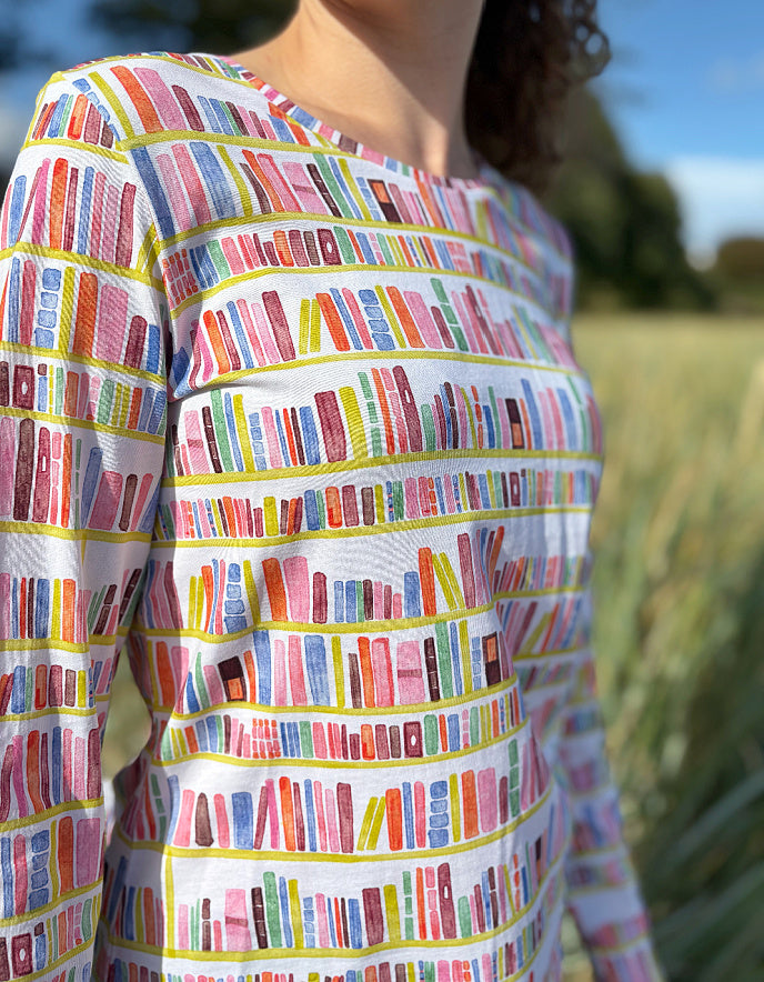 Woman wearing a colourful patterned long sleeve t shirt with bookshelf  pattern standing on a beach with grass and water in the background