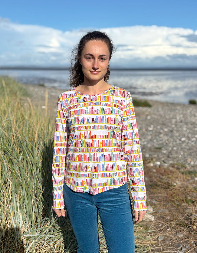 Woman wearing a colourful patterned long sleeve t shirt with bookshelf  pattern standing on a beach with grass and water in the background