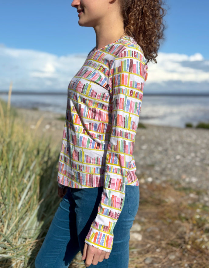 Woman wearing a colourful patterned long sleeve t shirt with bookshelf  pattern standing on a beach with grass and water in the background