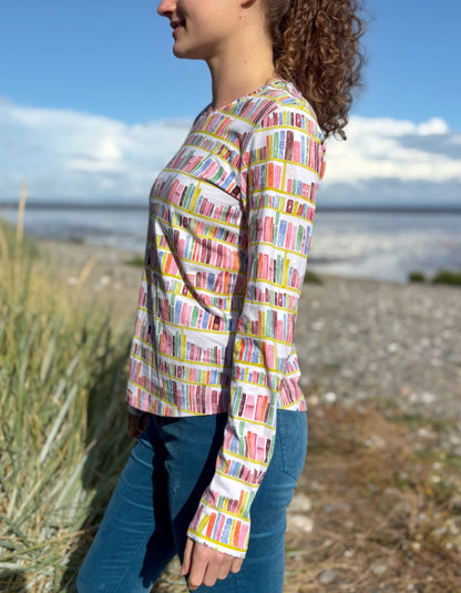 Woman wearing a colourful patterned long sleeve t shirt with bookshelf  pattern standing on a beach with grass and water in the background