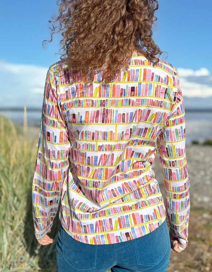 Woman wearing a colourful patterned long sleeve t shirt with bookshelf  pattern standing on a beach with grass and water in the background