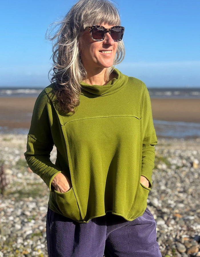 A woman with gray hair and sunglasses wearing an olive green long-sleeve cowl-neck top with pockets, standing on a pebble beach with the sea and blue sky in the background.