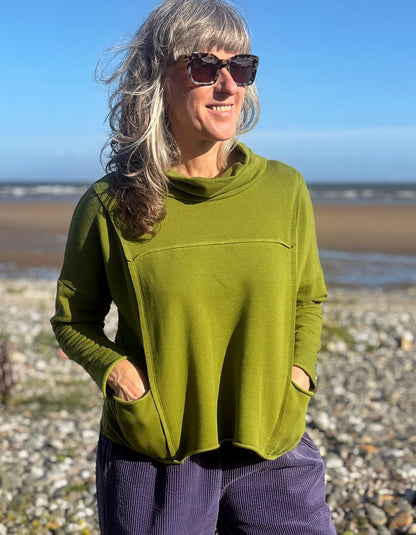 A woman with gray hair and sunglasses wearing an olive green long-sleeve cowl-neck top with pockets, standing on a pebble beach with the sea and blue sky in the background.