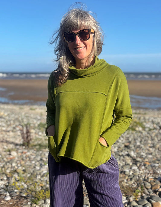 A woman with gray hair and sunglasses wearing a loose olive green cowl-neck top with side pockets and purple corduroy pants, standing on a pebble beach under a blue sky.