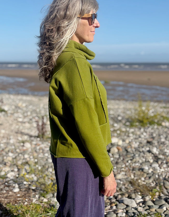 A woman with gray hair and sunglasses stands in profile on a rocky beach, wearing an olive green cropped fleece jacket and a purple ribbed skirt.