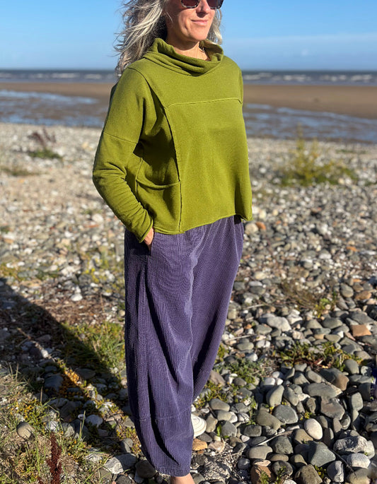A woman standing on a rocky beach wearing a loose olive green cowl-neck top and purple corduroy pants.