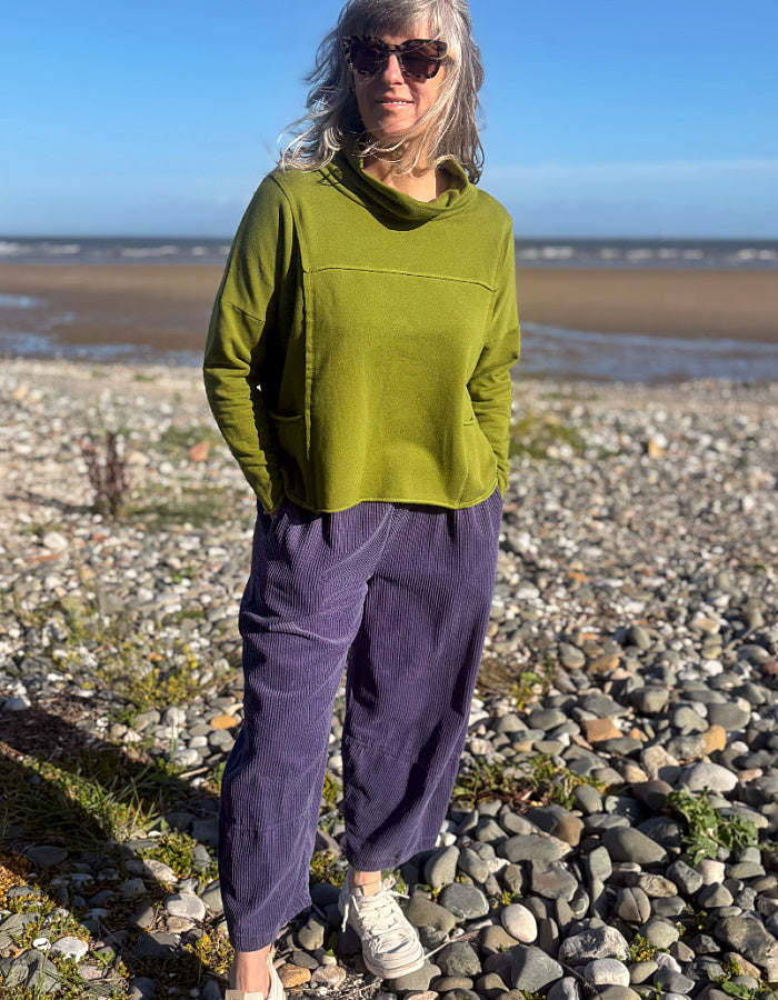 A woman standing on a rocky beach wearing a loose olive green long-sleeve top, purple corduroy pants, and white sneakers.