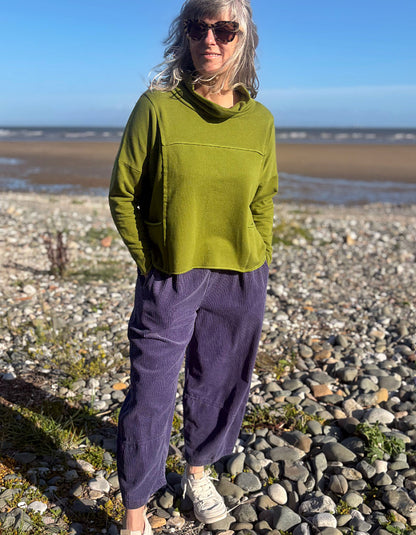 A woman standing on a rocky beach wearing a loose olive green long-sleeve top, purple corduroy pants, and white sneakers.