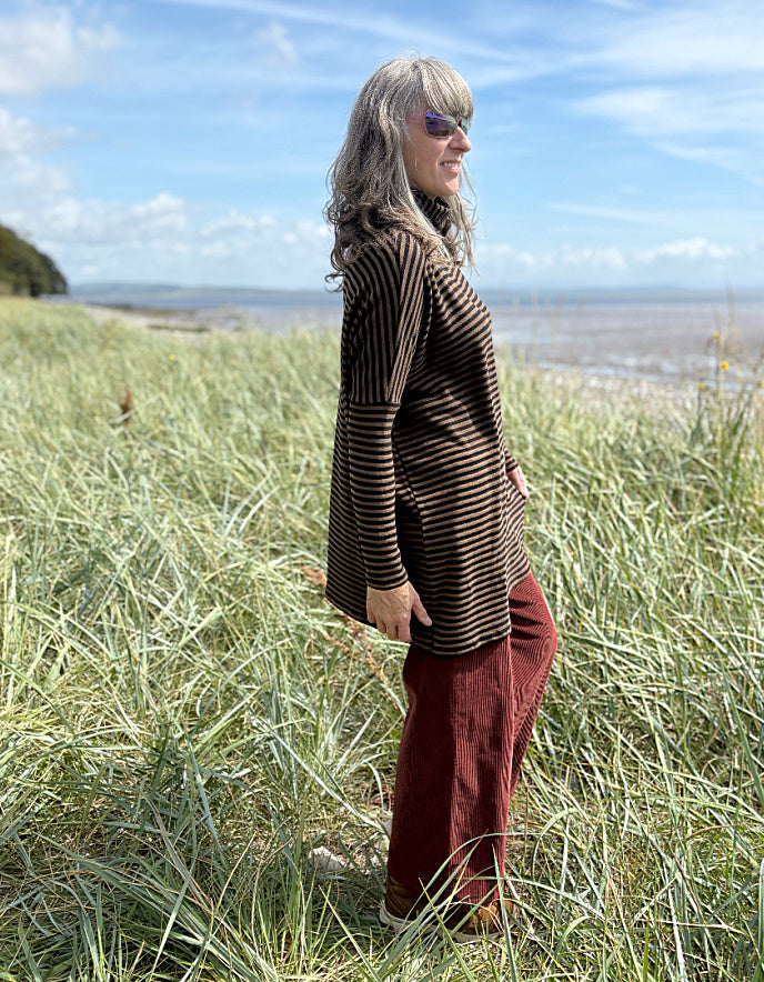 Person wearing a striped brown and black tunic and red pants standing in a grassy field with a blue sky and ocean in the background.