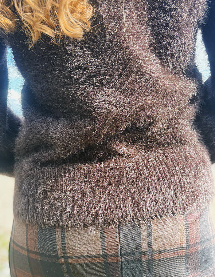 Close-up of a person wearing a fuzzy brown knit sweater and a plaid skirt in muted green, brown, and beige tones.