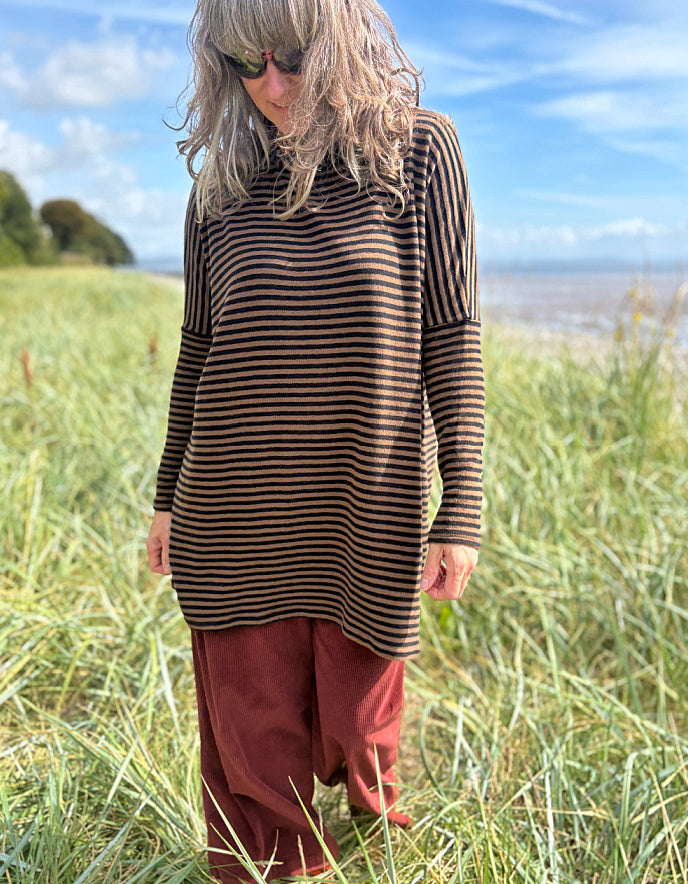 Person wearing a striped brown and black tunic and red pants standing in a grassy field with a blue sky and ocean in the background.