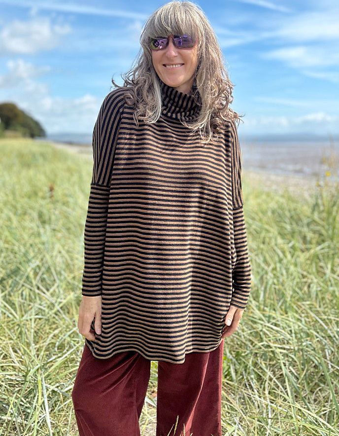 Person wearing a striped brown and black tunic and red pants standing in a grassy field with a blue sky and ocean in the background.