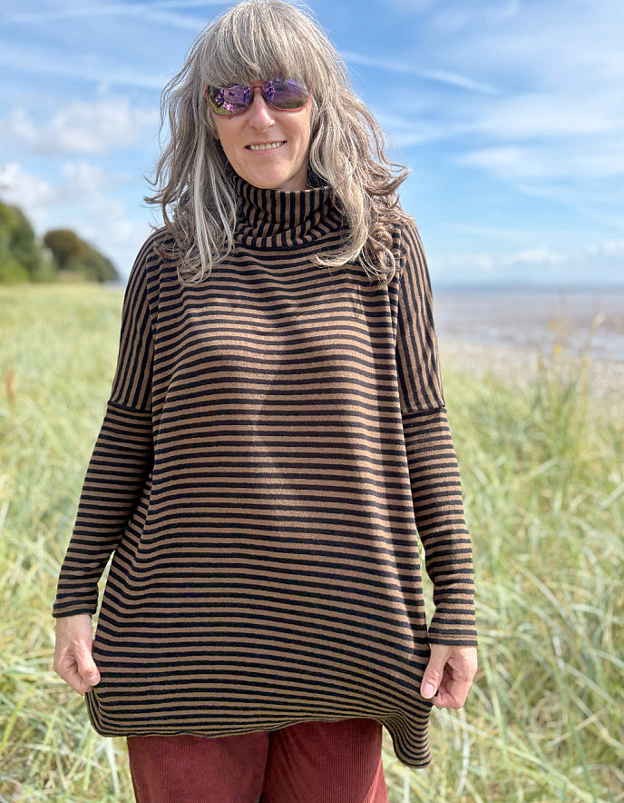 Person wearing a striped brown and black tunic and red pants standing in a grassy field with a blue sky and ocean in the background.