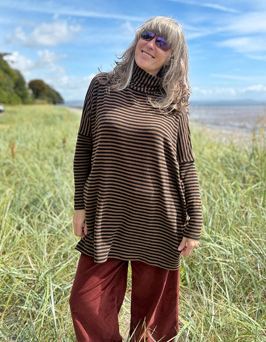 Person wearing a striped brown and black tunic and red pants standing in a grassy field with a blue sky and ocean in the background.