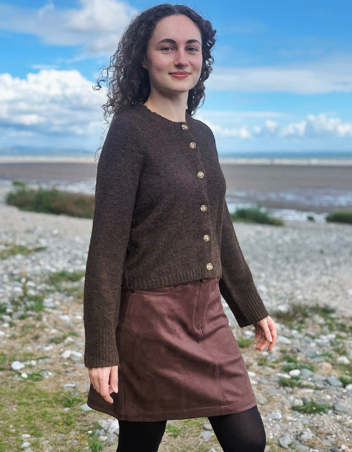 A woman standing on a rocky beach wearing a dark brown button-front cardigan and a brown skirt, with a blue sky and sea in the background.
