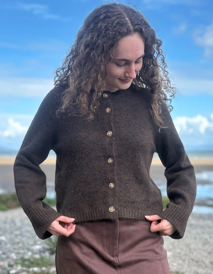 A woman with long curly brown hair wearing a dark brown button-up knit sweater and brown trousers, standing outdoors on a pebble beach with a blue sky and sea in the background.
