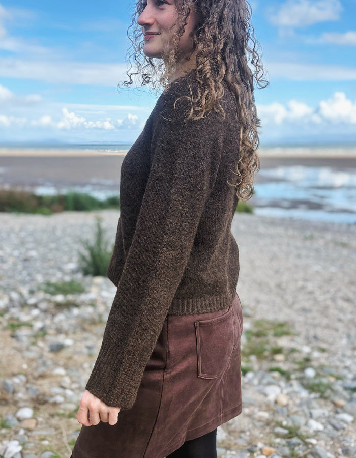 A woman with curly light brown hair stands on a pebble beach wearing a dark brown knit sweater and a brown skirt, with water and a cloudy sky in the background.