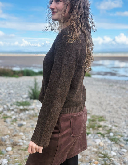 A woman with curly light brown hair stands on a pebble beach wearing a dark brown knit sweater and a brown skirt, with water and a cloudy sky in the background.