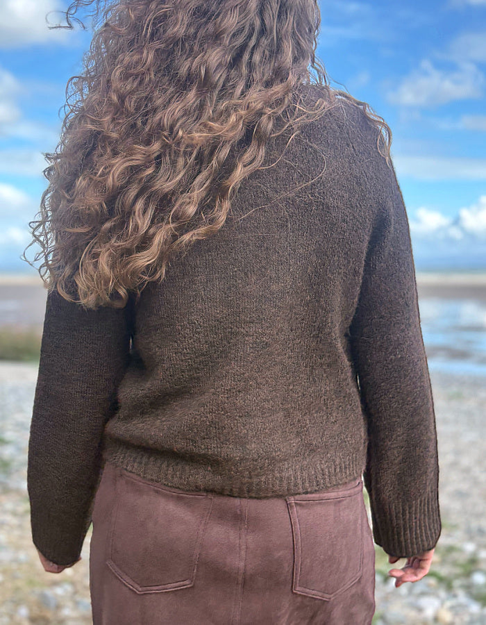 A person viewed from behind wearing a dark brown textured knit sweater and a dusty rose skirt, standing outdoors near a body of water under a blue sky.
