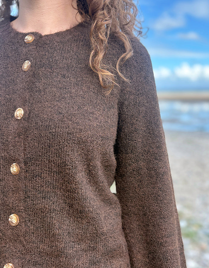 A close-up of a person wearing a brown knit sweater with large gold buttons, photographed outdoors with a blurred beach and blue sky background.