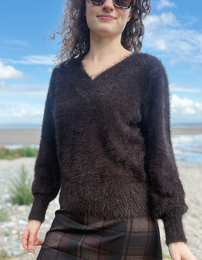 A woman wearing a dark brown fuzzy long-sleeve V-neck sweater and a brown plaid skirt, standing outdoors against a blue sky and water background.