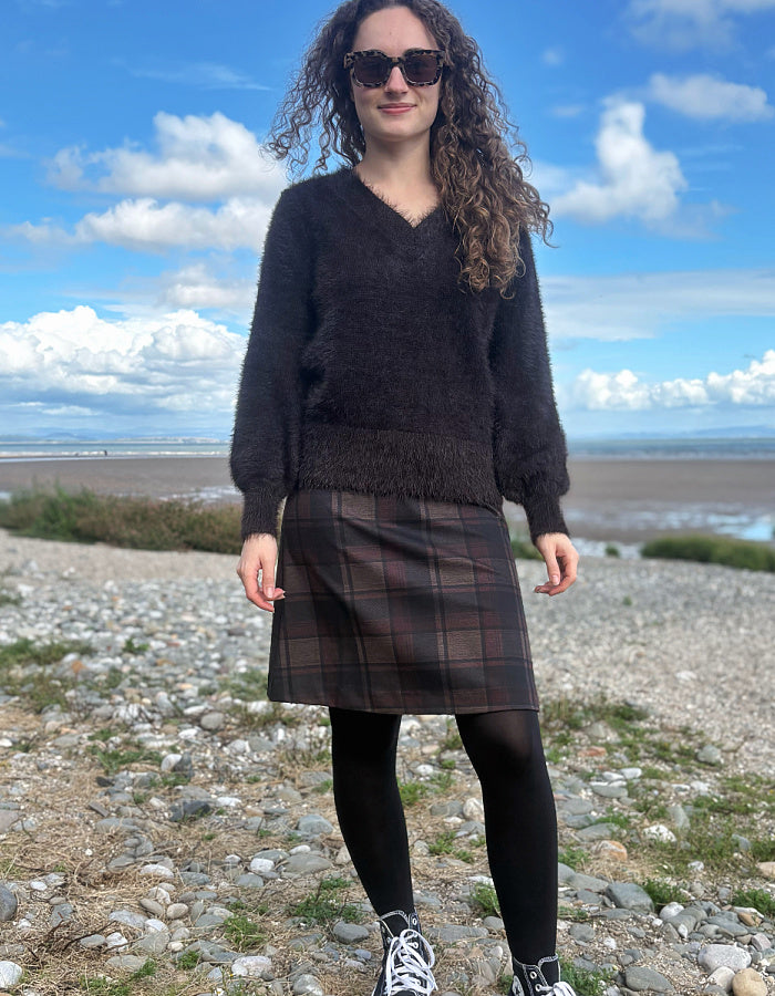A woman stands outdoors on a rocky shoreline under a blue sky with clouds, wearing a dark fuzzy V-neck sweater, a brown plaid skirt, black tights, and sneakers.