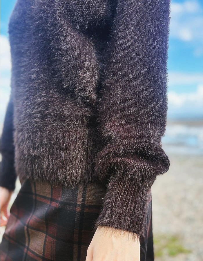 A close-up of a person outdoors wearing a fuzzy dark brown or charcoal knit sweater with a plaid skirt, with a blurred beach and blue sky background.