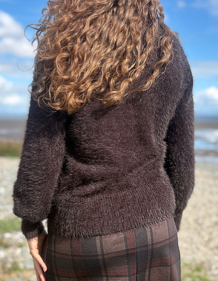 A person viewed from behind wearing a dark brown fuzzy knit cardigan and a brown plaid skirt, standing outdoors on a pebble beach with blue sky in the background.