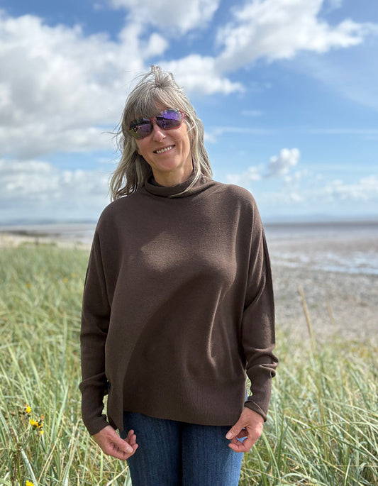 Person wearing a brown sweater with mock turtle neck standing in grass with a beach and blue sky in the background