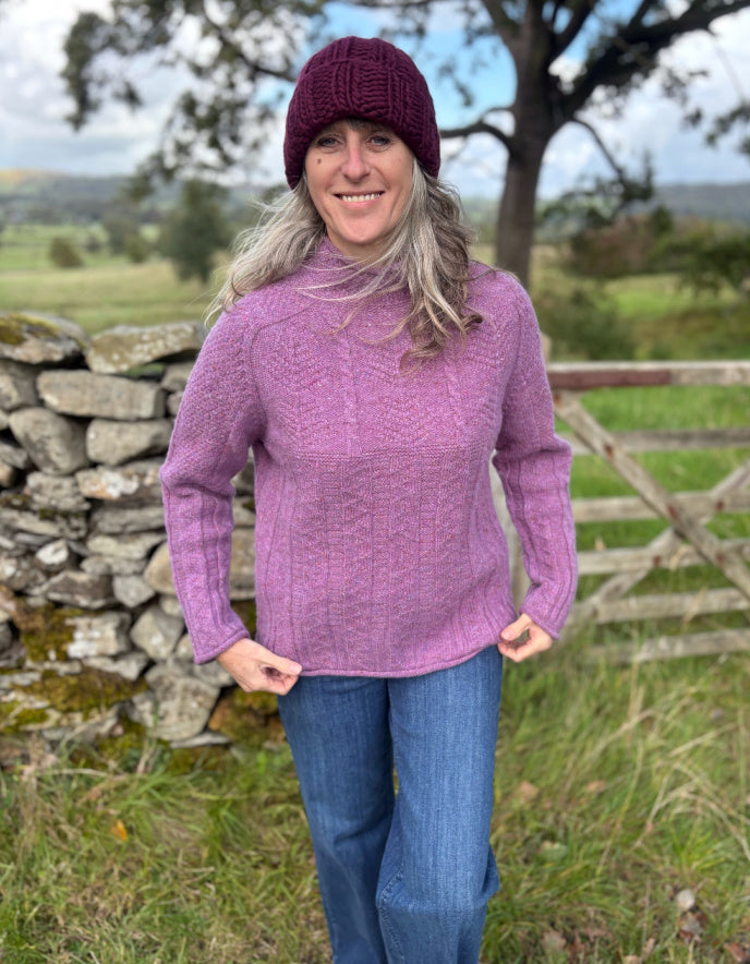 Person wearing a purple sweater and hat standing in a field with a stone wall and trees in the background.