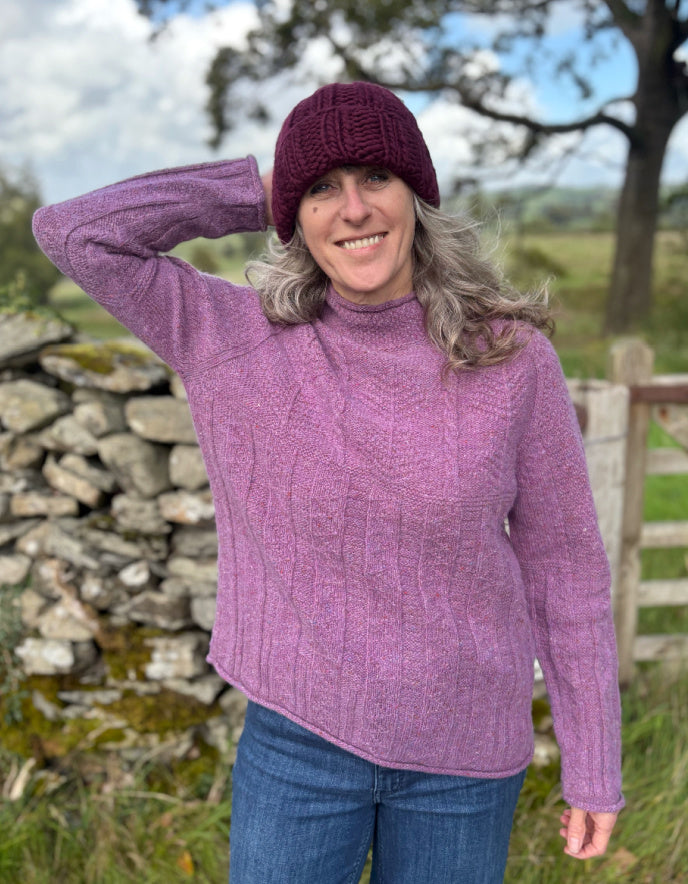 Woman wearing a purple cable tweed sweater and hat outdoors with a stone wall and tree in the background