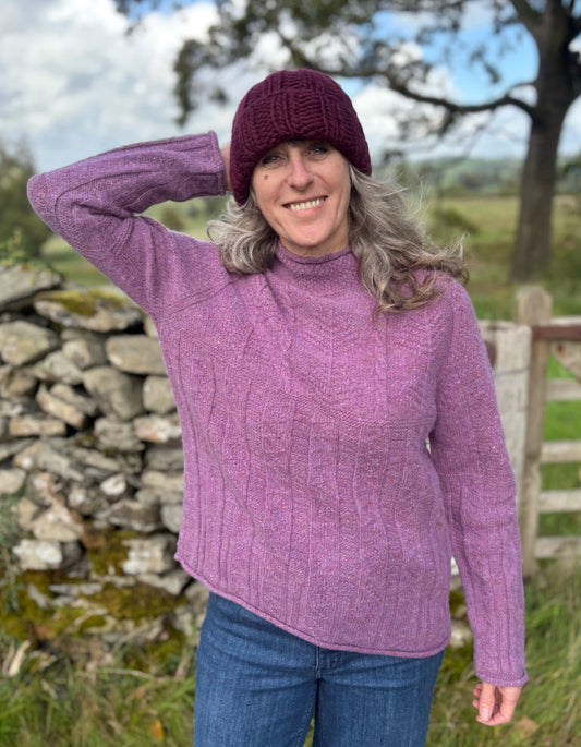 Woman wearing a purple cable tweed sweater and hat outdoors with a stone wall and tree in the background