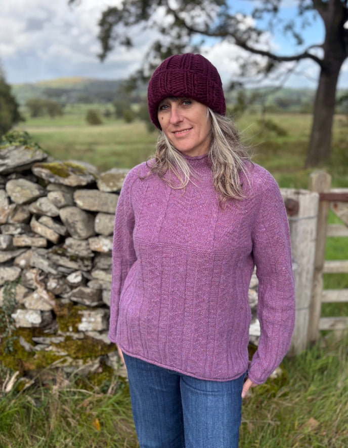Woman wearing a purple cable tweed sweater and hat outdoors with a stone wall and tree in the background