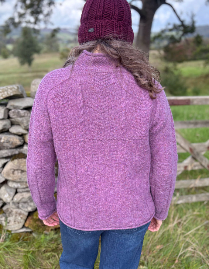 Woman wearing a purple cable tweed sweater and hat outdoors with a stone wall and tree in the background