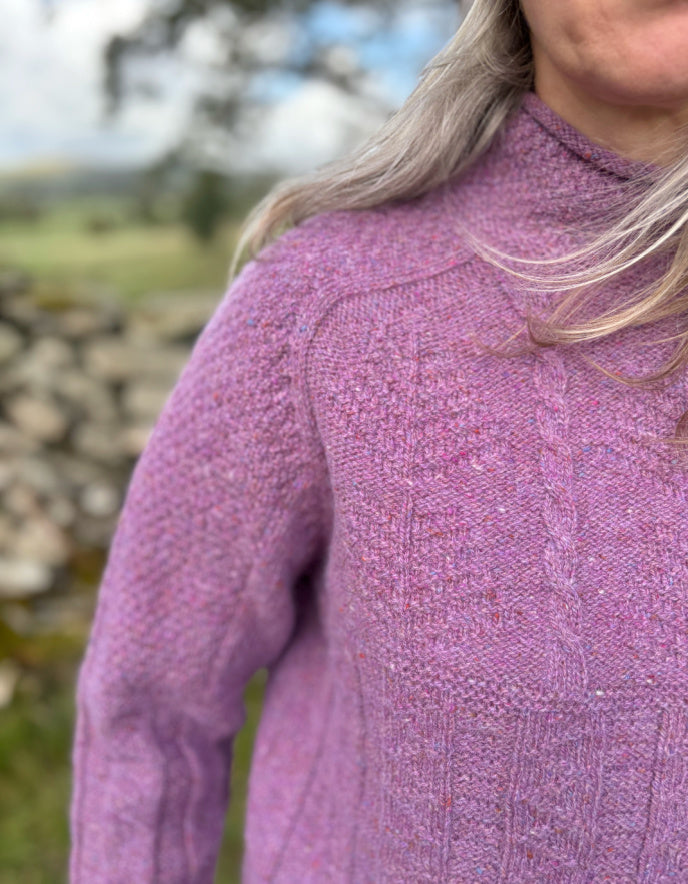Woman wearing a purple cable tweed sweater and hat outdoors with a stone wall and tree in the background