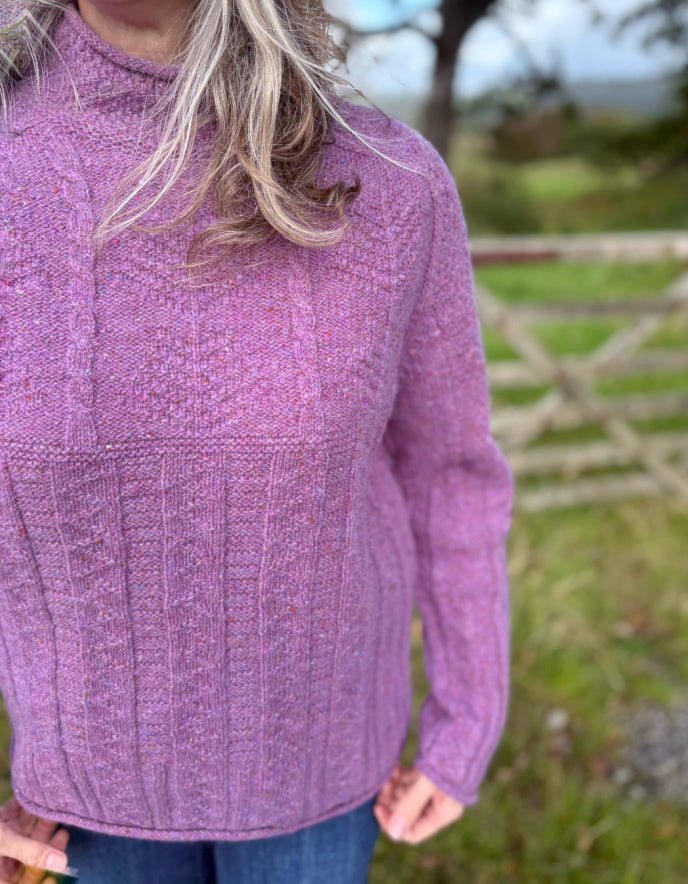 Woman wearing a purple cable tweed sweater and hat outdoors with a stone wall and tree in the background