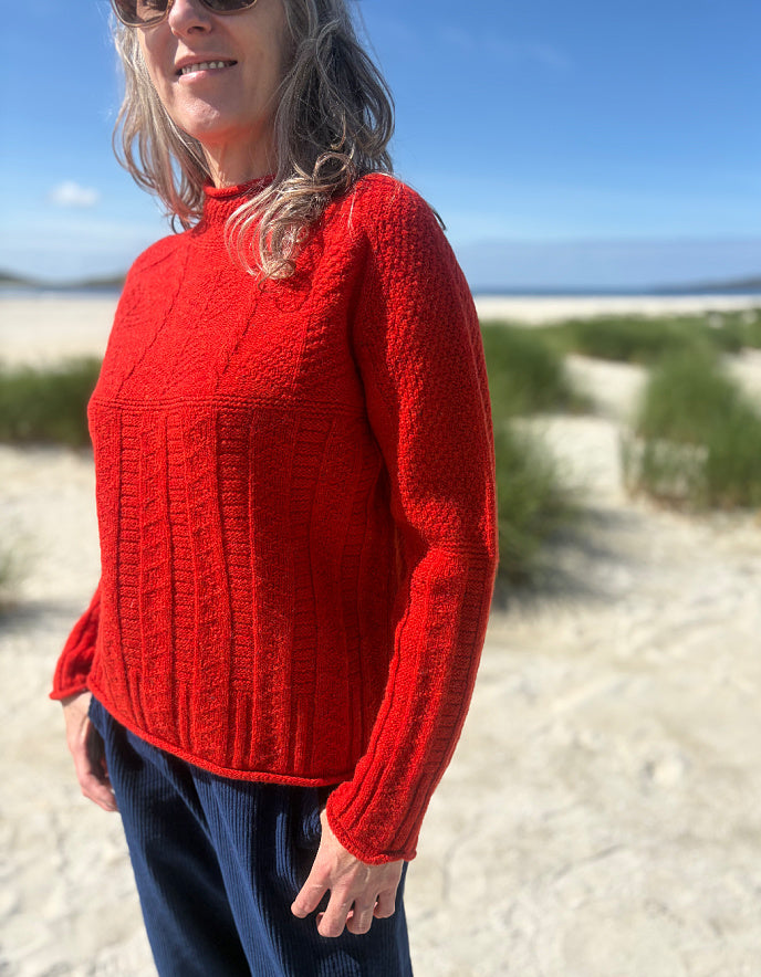 A woman wearing a bright red cable-knit sweater and navy pants, standing outdoors on a sandy beach with dunes and blue sky in the background.