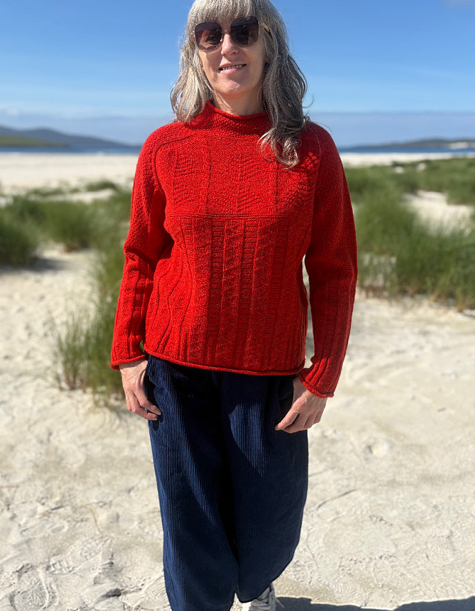 A woman standing on a sandy beach wearing a bright red cable-knit mock-neck sweater and dark blue pants.