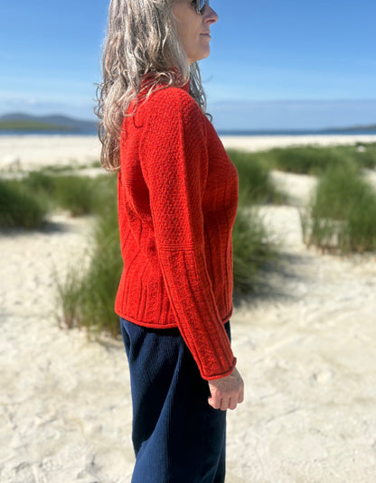 A woman standing on a sandy beach wearing a bright red cable-knit sweater and dark navy pants, shown in profile against a background of dunes, ocean, and blue sky.