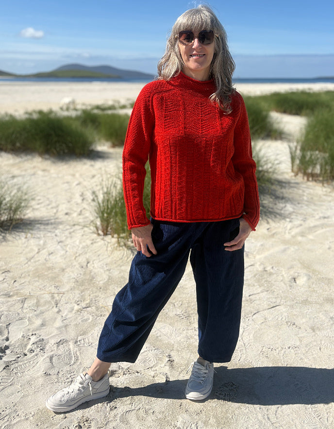 A woman stands on a sandy beach wearing a bright red cable-knit mock-neck sweater, dark navy wide-leg pants, and white sneakers.