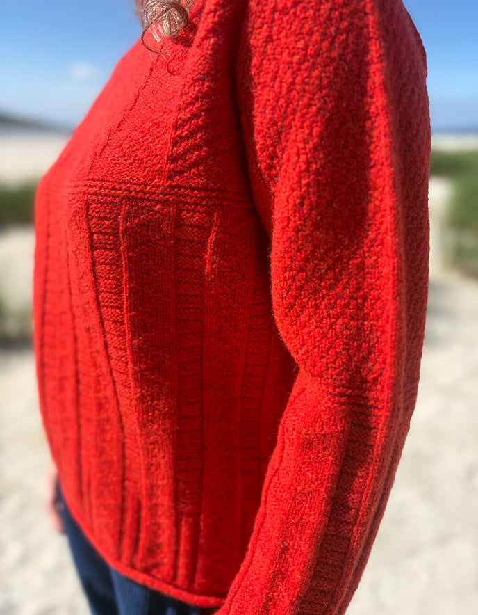 A close-up side view of a person wearing a bright red textured knit sweater outdoors on a sandy beach with blurred greenery and sky in the background.